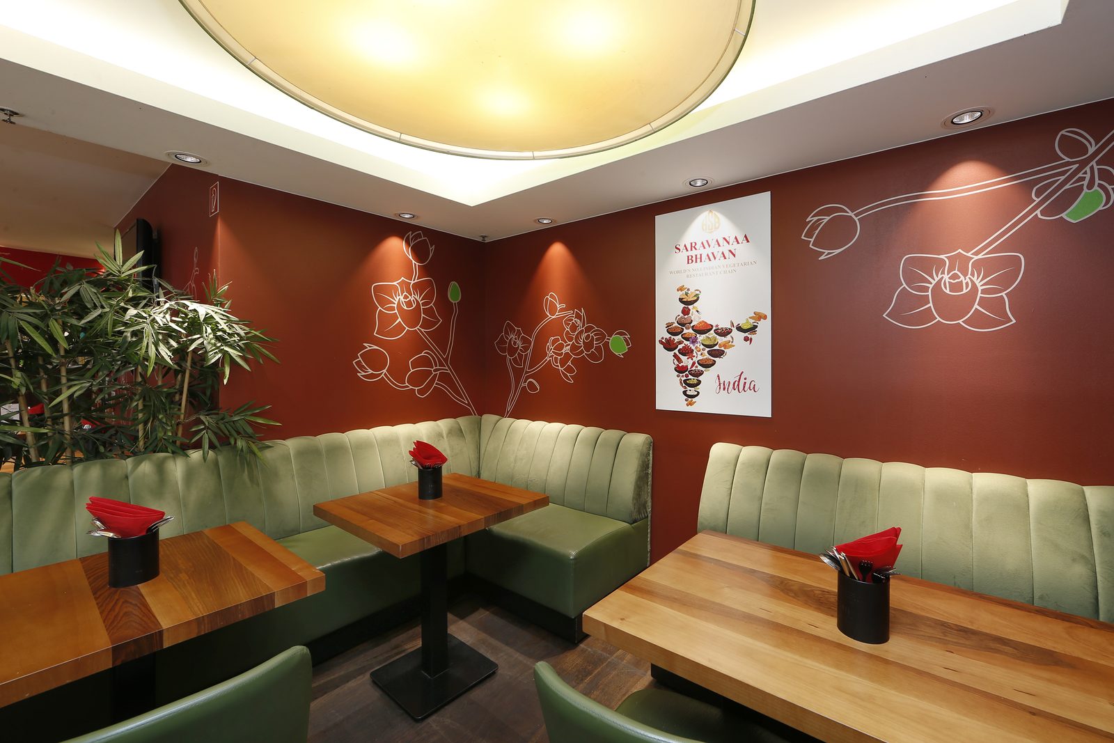 Corner with green upholstered booths, wooden tables and red napkins; a Saravanaa Bhavan poster on the wall.