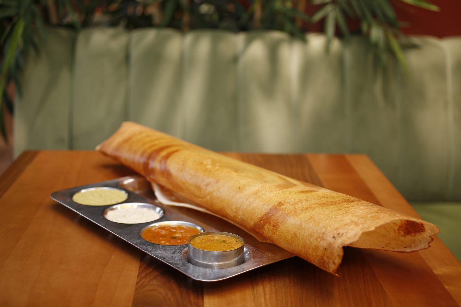 Lange, goldbraun gerollte Dosa auf einem Stahltablett mit vier Schälchen Chutney und Sambar.