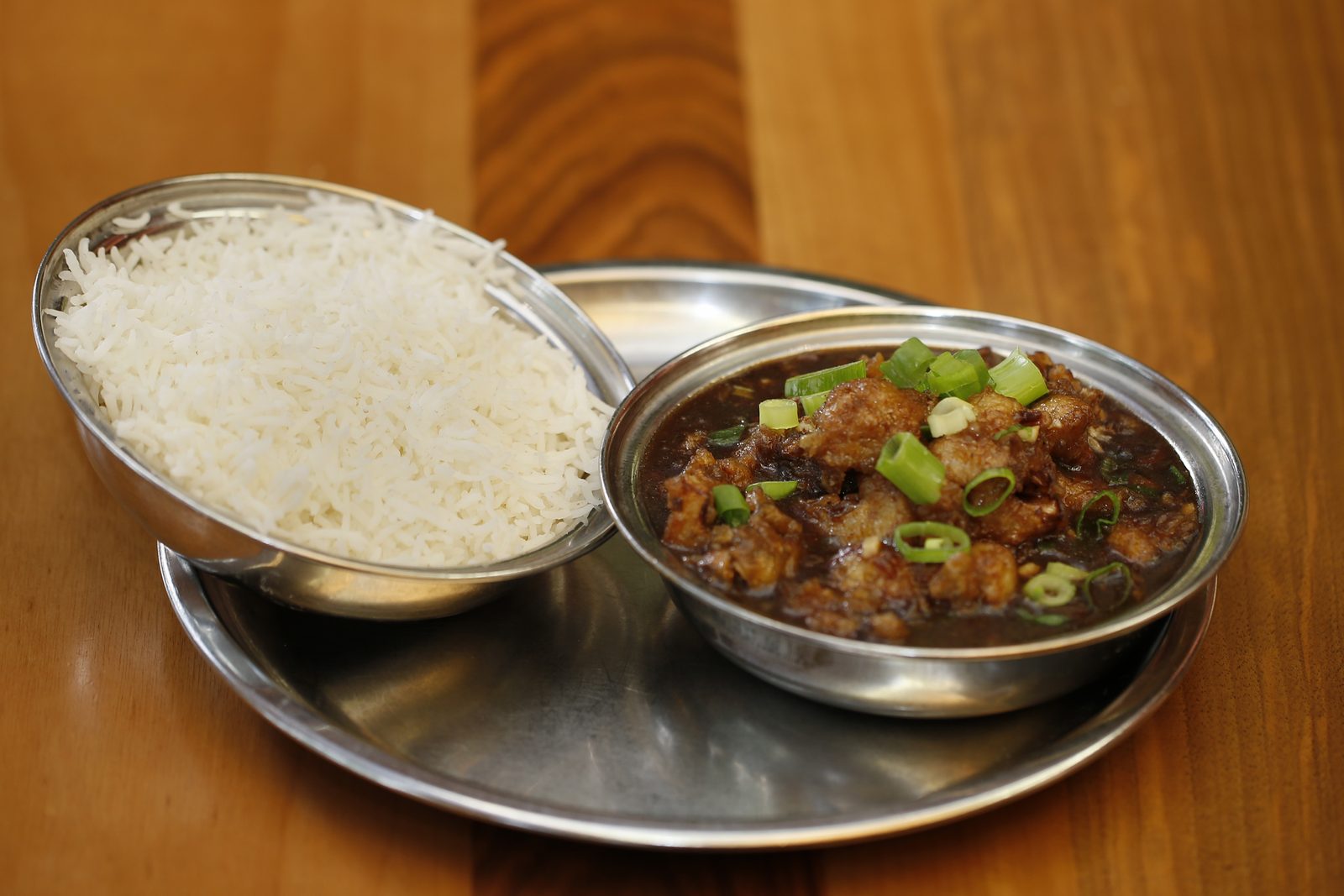 Manchurian in dunkler Sauce mit Frühlingszwiebeln, daneben eine Schale Basmatireis.